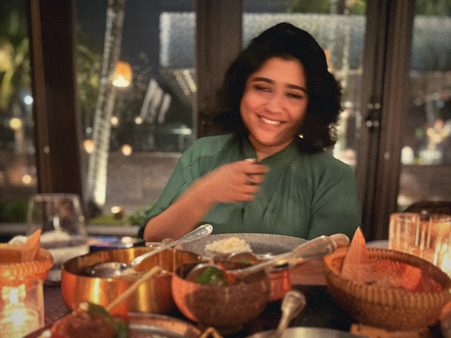 Divya laughing at a candlelit table spread with curries and copper bowls