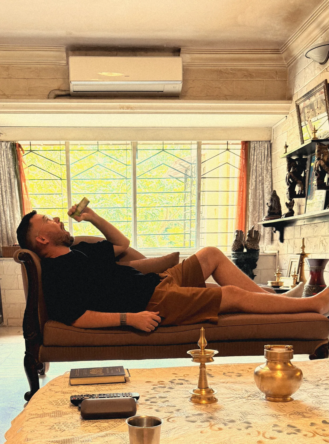 Steven stretched out on a carved daybed with a snack, a brass lamp on the coffee table below