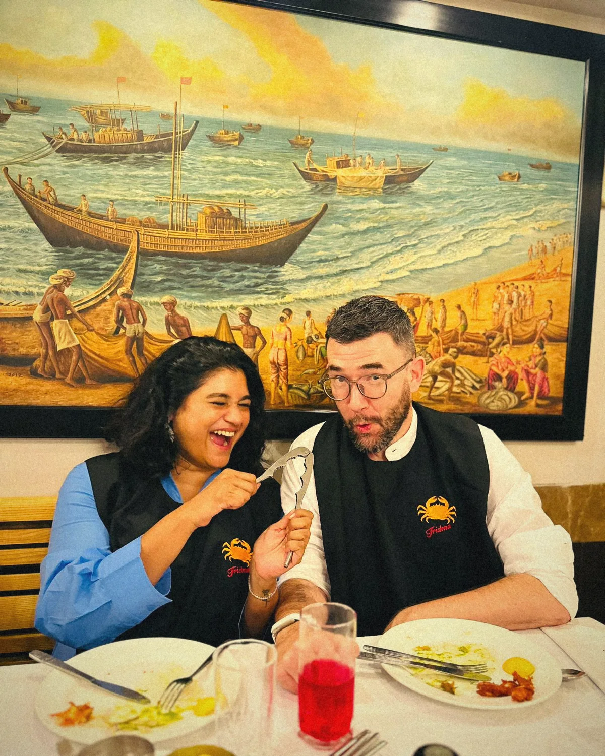 Divya laughing and holding a crab cracker next to Steven, both in Trishna crab bibs, under a painting of fishing boats